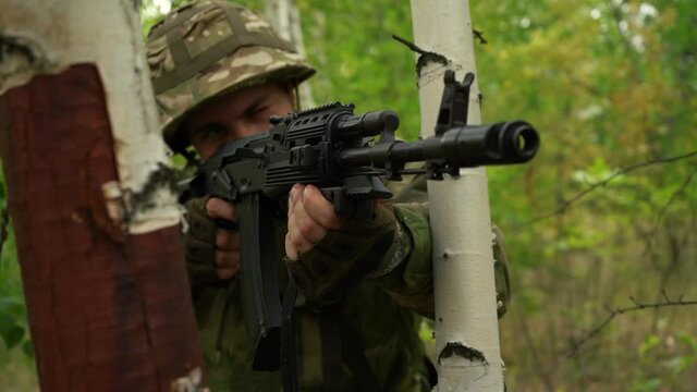 soldier aiming with a machine gun, looking out from behind a tree in the forest