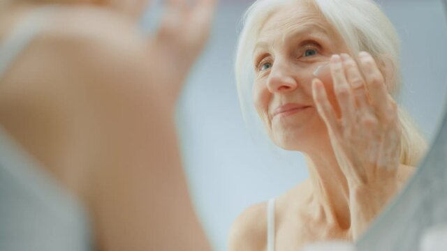 Portrait of Beautiful Senior Woman Morning Routine, Looking into Mirror Gently Applying Face Cream. Elderly Lady Makes Her Skin Soft, Smooth, Wrinkle Free with Natural anti-aging Cosmetics, Products
