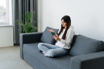 Indignant young woman sitting on couch in living room holding mobile phone having problems with digital device, feels angry and annoyed.