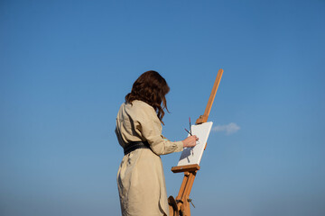 Female Painter artist paints a picture on white empty canvas outdoors blue sky