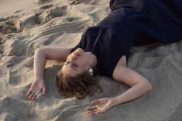 Beautiful woman in a black dress lies on the sand near the sea at sunset