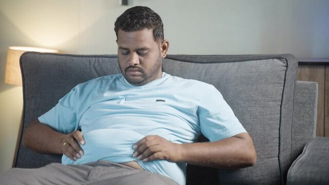 Sad obese man disappointed for heavy belly fat by holding hands while sitting on sofa at home - Concept of overweight, disease, suffering from excess stomach waist and unhealthy eating problem.