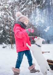 Little girl in a bright jacket plays in the winter snowy forest with her dog