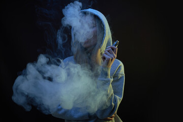 Portrait of beautiful blonde girl in gray hoodie smokes vape isolated on black studio background,...