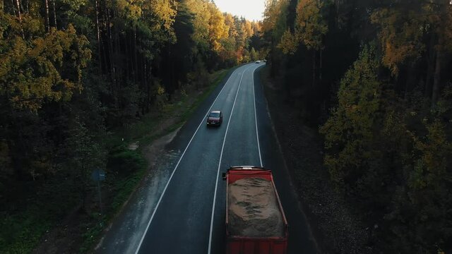 Automobiles and lorries drive along regional asphalt road stretching through autumn forest with colorful leaves on trees close aerial view