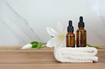 Two bottles of cosmetics with an eyedropper on a background of white marble tiles. In the background are folded towels and white flower. The concept of spa skin care and relaxation with essential oil.