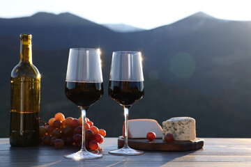 Red wine served with cheese and grapes on wooden table against mountain landscape