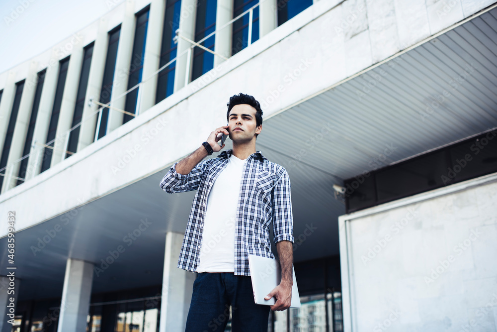 Poster caucasian man with modern laptop computer making consultancy cellphone conversation during daytime n