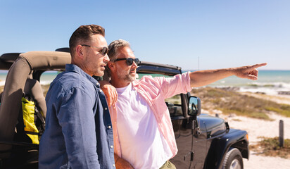 Happy caucasian gay male couple embracing and pointing by a car at seaside