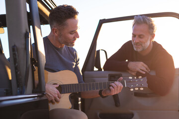 Happy caucasian gay male couple playing guitar and smiling, hanging out by car at seaside