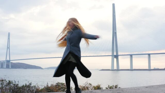 Zooming out aerial view of a smiling woman with long hair dancing against the cable-stayed Russian bridge in Vladivostok, Russia. Cloudy morning