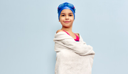 A positive little girl in a swimsuit, goggles, and blue swimming cap looking to the camera covering her wet body with a white towel after swimming, isolated blue studio background.