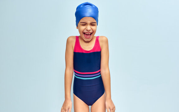 Studio image of a little funny happy girl in a swimsuit, isolated over blue studio background. A joyful wet kid in a swimsuit and swim cap smiling broadly during swimming training.