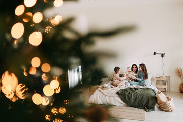 Fototapeta na wymiar A happy mom with two daughters is resting, relaxing and having fun sitting on the bed in a cozy decorated bedroom during the Christmas holiday at home during the New Year vacations. Selective focus