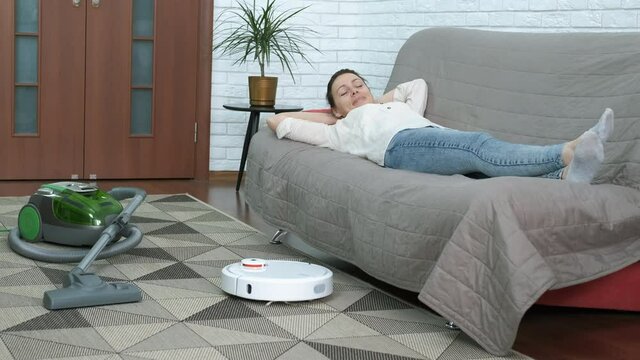 Relax with housekeeping. A housewife lay on the sofa while a white round vacuum cleaner wipe the dust on the floor.