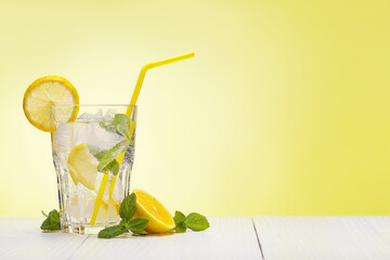 Glass of water with fresh lemons, leaves of mint and ice cubes on table against yellow background,...