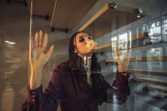Young woman behind a glass wall with a flower in her mouth. Creative portrait.