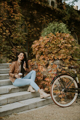 Young woman with mobile phone drink coffee to go at the stairs by the bicycle on autumn day