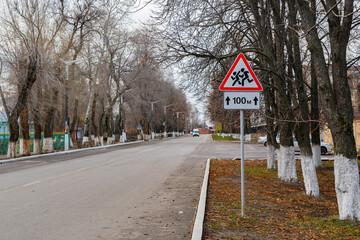 Road sign "beware of children", next to the educational institution. The road sign is a white triangle, with black running figures and red edging.