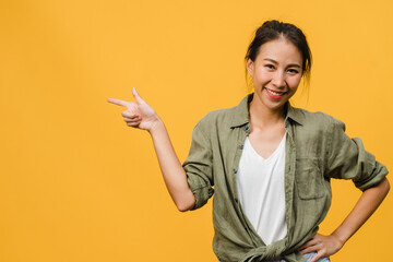 Portrait of young Asian lady smiling with cheerful expression, shows something amazing at blank space in casual cloth and looking at camera isolated over yellow background. Facial expression concept.