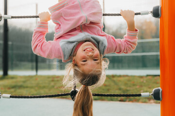A young girl on a climbing frame hangs upside down on a children's playground.