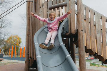A little cheerful girl with long blond hair goes down the children's slide.