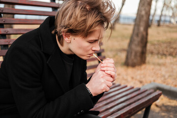 Young man with birthmarks on face in black coat sitting on wooden bench and lighting up a cigarette...
