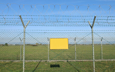 Empty yellow sign outside a double wire mesh barrier with barbed wire on top. Background for copy space
