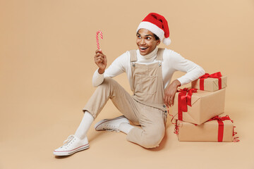 Full size young african man wear Santa Claus red Christmas hat sit near many present gifts boxes eat candy cane lollipop isolated on plain pastel beige background studio. Happy New Year 2022 concept.