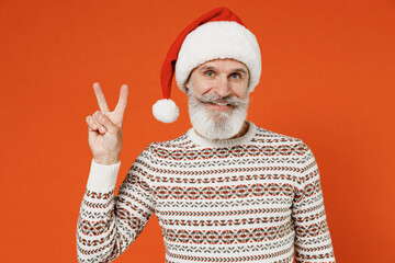 Old happy bearded Santa Claus man 50s wear Christmas hat sweater posing showing victory sign isolated on plain orange background studio. Happy New Year 2022 celebration merry ho x-mas holiday concept