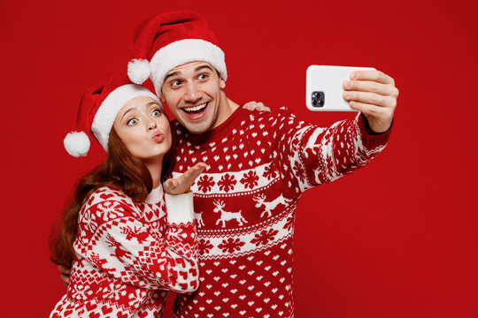 Young couple friends two man woman in sweater hat doing selfie shot on mobile cell phone blow air kiss isolated on plain red background. Happy New Year 2022 celebration merry ho x-mas holiday concept.