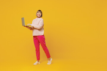 Full body elderly smiling happy caucasian copywriter woman 50s wears pink casual knitted sweater hold use work on laptop pc computer look aside isolated on plain yellow background studio portrait