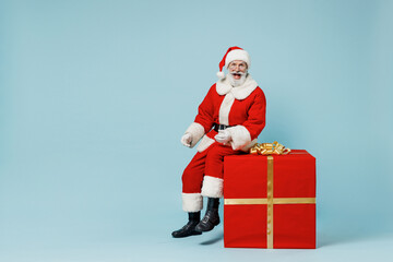 Full size old Santa Claus man in Christmas hat red suit clothes sitting on big gift present box isolated on plain blue background studio Happy New Year 2022 celebration merry ho x-mas holiday concept