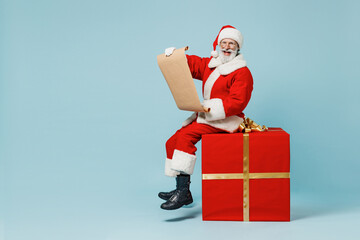Full size fun old Santa Claus man in Christmas hat red suit clothes sit on big gift present box read letter isolated on plain blue background studio Happy New Year 2022 merry ho x-mas holiday concept.