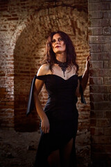 A brunette girl with long hair in an evening dress in a vintage Gothic castle. A woman who looks like an evil witch or sorceress in an abandoned old church. Female Model posing in Halloween