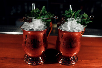 two cocktails with ice in copper mugs with ice decorated with pine cones and mint on a wooden bar on a dark background