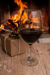 glass with red wine next to a gift, background a fireplace with fire, cozy place with Christmas decoration, drink