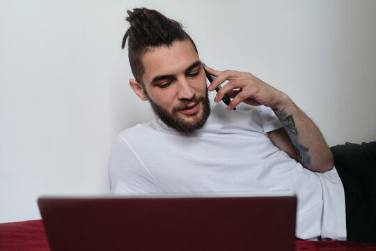 Conduct business via Internet online. Young handsome freelancer man in white Tshirt with beard and dreadlocks works at home on self isolation. Talk on phone and discuss important matters.