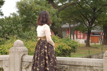 Mujer en un hanbok parada en un puente de piedra mirando el Palacio Gyeongbokgung rodeado por el bosque - obrazy, fototapety, plakaty