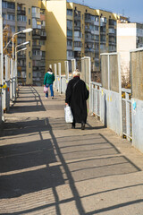 An old woman in a black coat walks across an old bridge following a young woman at a safe distance towards city houses under a blue sky