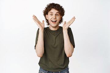 Portrait of excited young man with curly hair, looking fascinated and surprised, clap hands and...