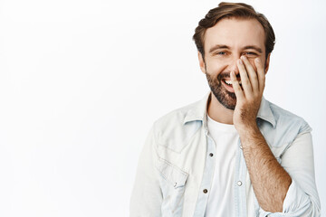 Image of happy adult man laughing and smiling, holding hand on his face and blushing carefree, white background