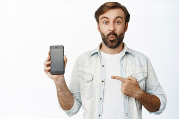 Surprised bearded man pointing at his cellphone screen, showing promo deal in app, standing over white background