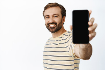 Portrait of handsome smiling casual guy in t-shirt, extending hand with phone, showing smartphone screen, demonstrating app interface, white background