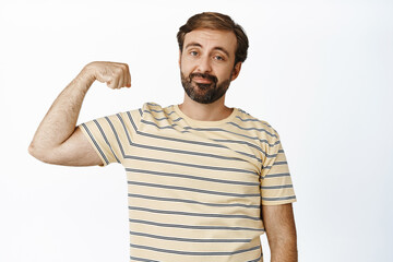 Funny bearded guy shows his biceps, flexing muscles on arms and looking at camera, standing in casual t-shirt over white background