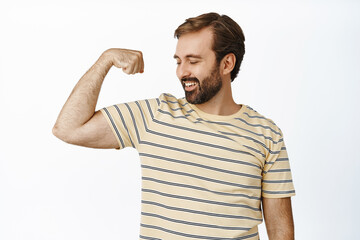 Portrait of smiling pleased man looking at his arm muscles, flexing biceps with satisfied face expression. Concept of workout and gym membership