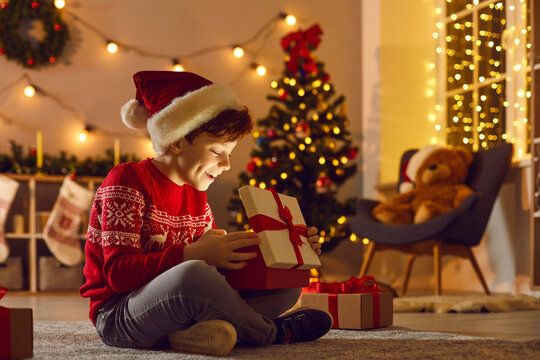 Just believe in magic. Happy surprised kid in Santa hat unpacks New Year gift. Luminous glow lights up little boy's face as he opens his wonderful present sitting in the living-room on Christmas night