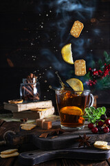 Tea with lemon, cinnamon and star anise on dark wooden boards. Cookie slices and lemon slices hang in the air. Splashes and clouds of steam around. A jar with cinnamon tubes in the background