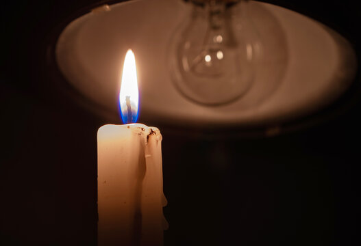 Burning candle near a switched off light bulb in complete darkness. Blackout, electricity off, energy crisis or power outage, concept image. 