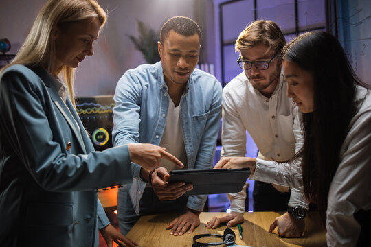 Four diverse colleagues standing at office room and looking together at digital tablet. Male and female company workers discussing business ideas during evening conference.